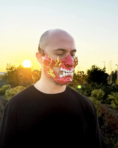 Man wearing Fire Red Gold Samurai Demon Oni red mask with gold teeth, handcrafted Venetian cosplay mask at sunset.