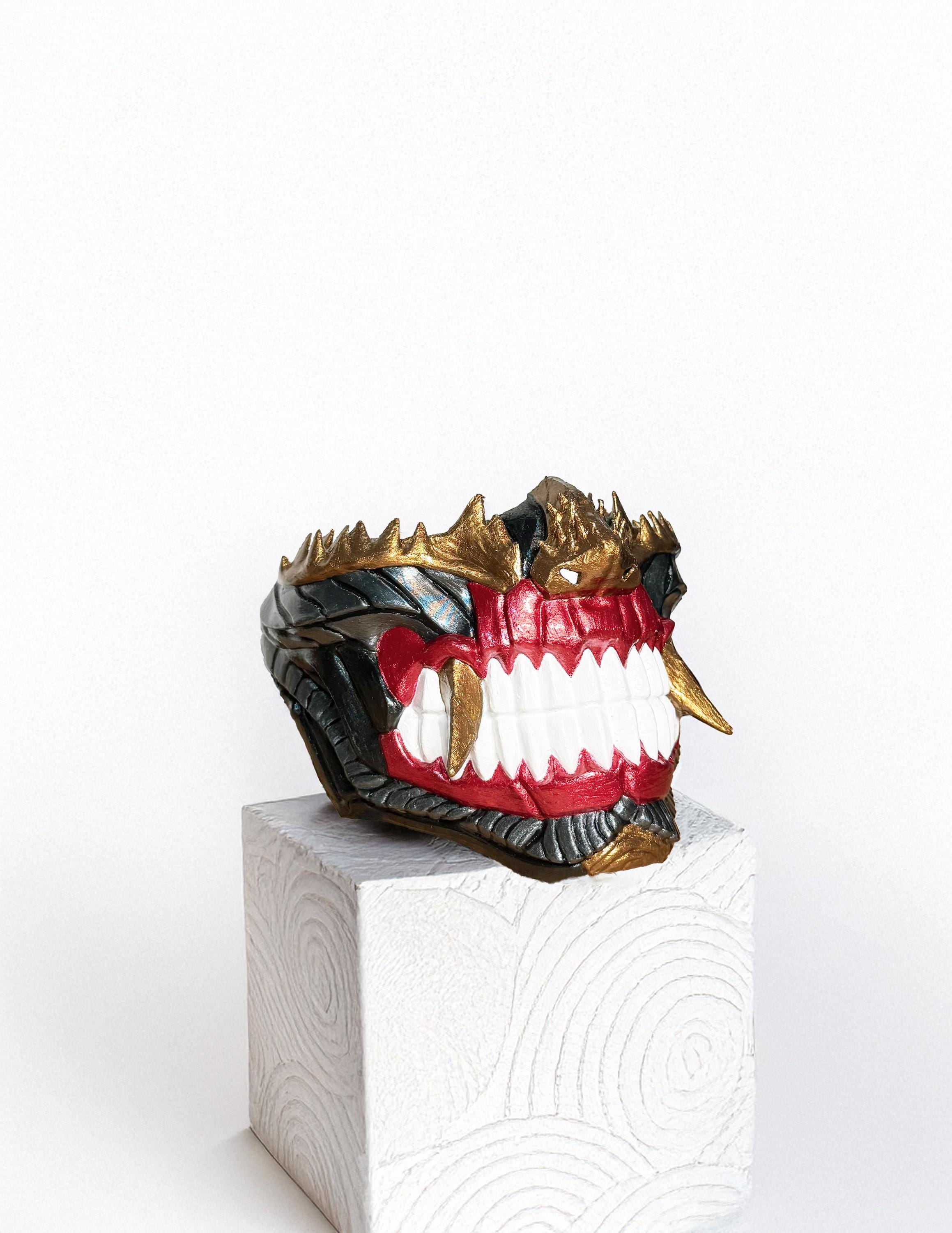 May include: A red, gold, and black mask with sharp teeth and a textured surface. The mask is sitting on a white pedestal with a swirling pattern.
