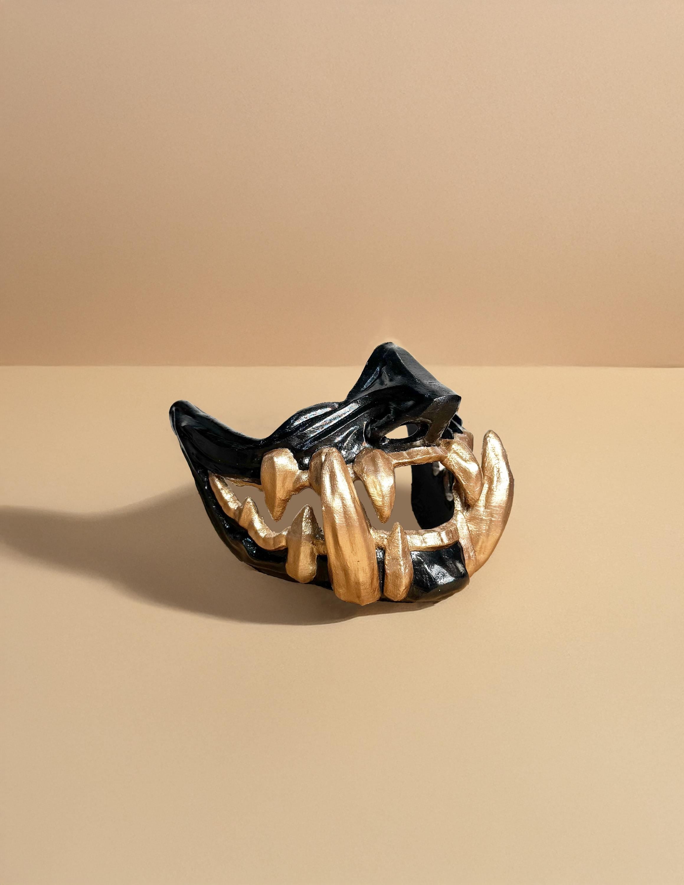 May include: A black and gold mask with a sharp, jagged design. The mask has a wide mouth with gold teeth and a black, textured surface.